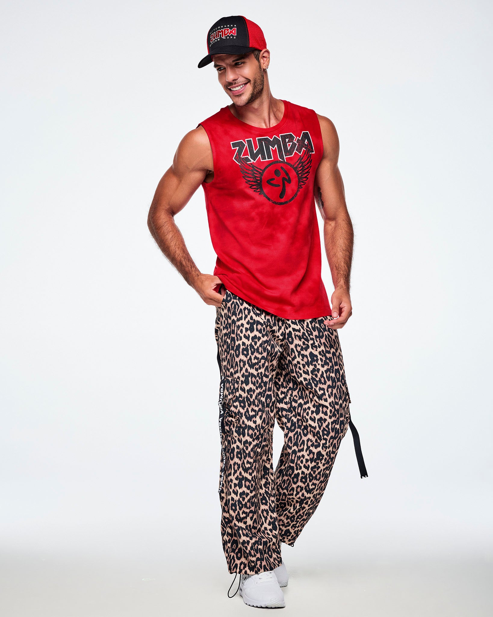 A male model wearing a black and red ZUMBA hat, red Zumba rock tank top and paired with leopard print cargo pants and white shoes.