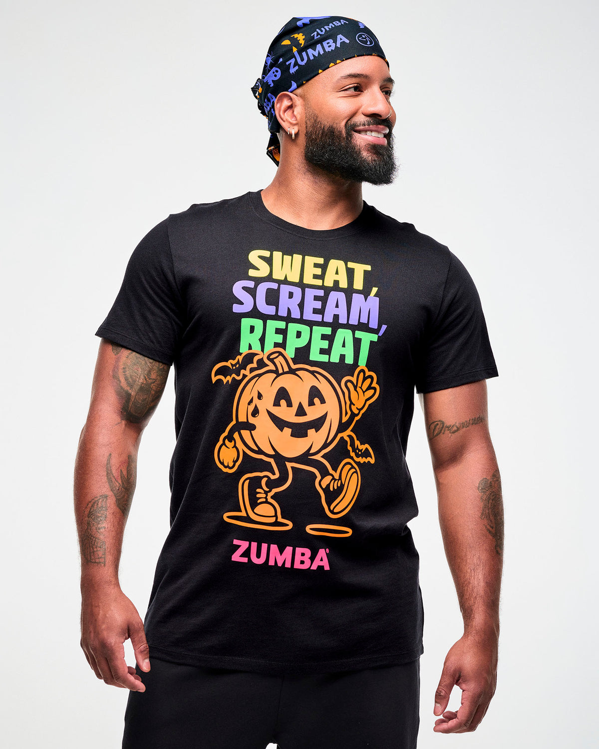 Male model wearing a black and purple Halloween bandana as a headwrap with a black "Sweat, Scream, Repeat" tee featuring a pumpkin graphic and "ZUMBA" in pink text.