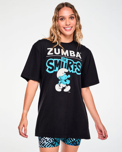Female model in a black crew neck tee with white 'ZUMBA' text over a line and a blue 'THE SMURFS' logo, above a walking Smurf in a white hat and pants. Paired with blue and black logo print biker shorts.