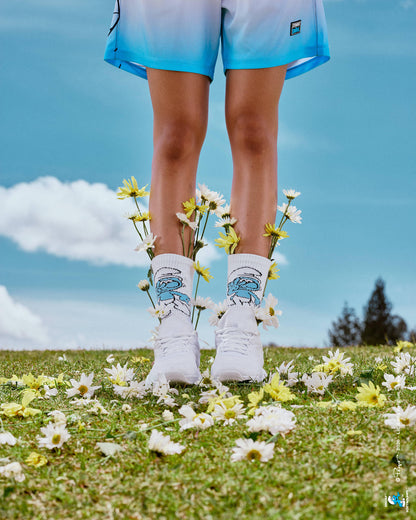 Close-up of a female model's legs in the Smurf crew socks and white shoes, with daisies tucked into the socks and scattered on the grass, and navy-to-light-blue ombre mesh shorts at the top of the frame, all against a blue cloudy sky.