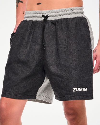 Male model wearing black denim and grey shorts with a drawstring waist, white "ZUMBA" text on the lower left hem, and white sneakers, hand in the side pocket.