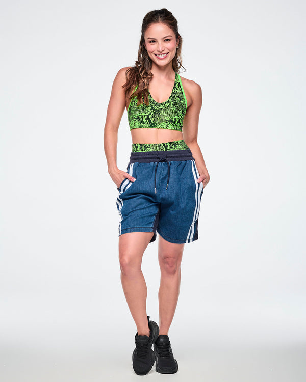 Female model wearing a neon green snake-print bra top with denim combo shorts featuring a navy back panel and double white stripe, paired with black sneakers.