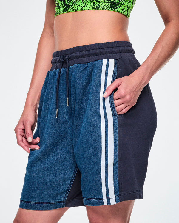 Close-up front view of a female model with her hands in the side pockets of denim combo shorts, showing the denim leg with a double white stripe beside the navy back panel and drawcord.