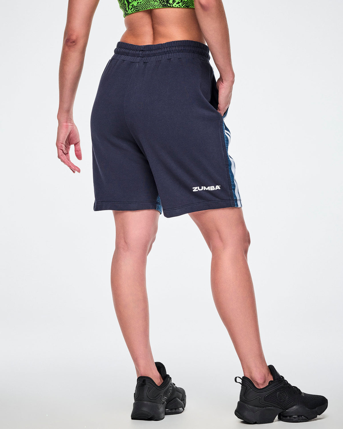 Back view of a female model wearing navy combo shorts with white "ZUMBA" text at the hip and a denim front panel partially visible. 