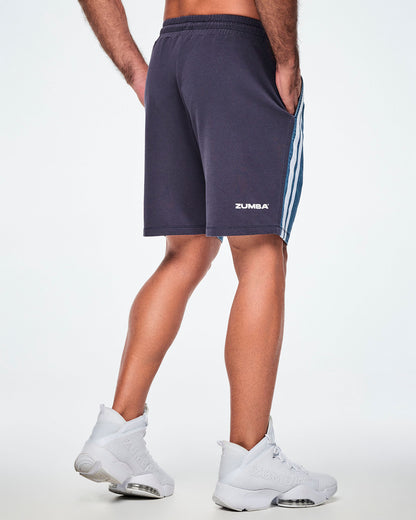 Back view of a male model wearing navy combo shorts with white "ZUMBA" text at the hip and a denim front panel seen from the side.