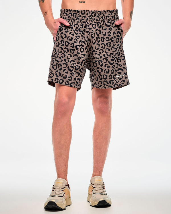 Male model in brown easy shorts with an all-over black leopard print, an elastic waistband, and white 'ZUMBA' text near the hem, both hands in the the side pockets.