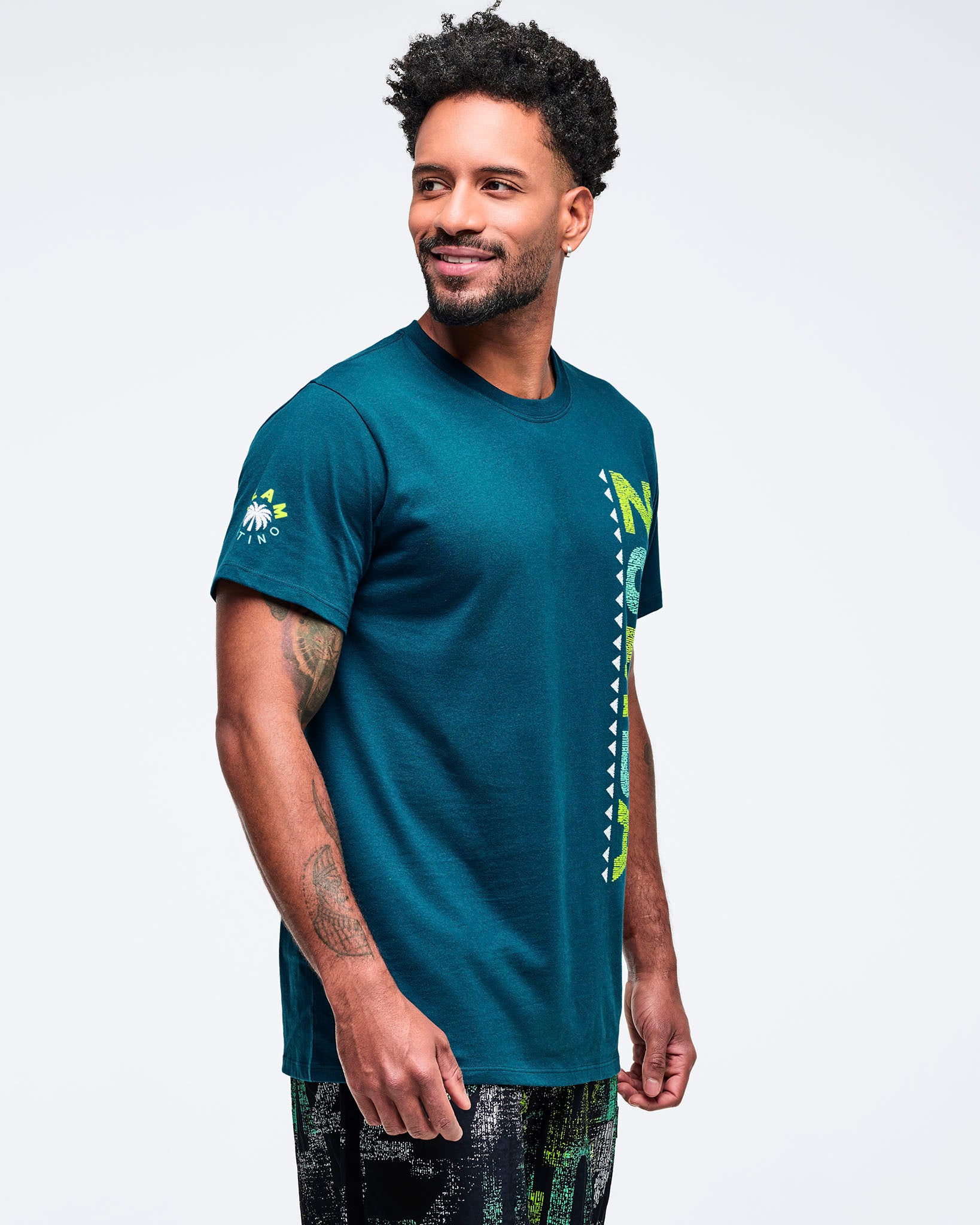 Teal crewneck T-shirt with short sleeves and a vertical lime “ZUMBA” graphic along the side front, shown in side view on a male model wearing patterned shorts on a white studio background.