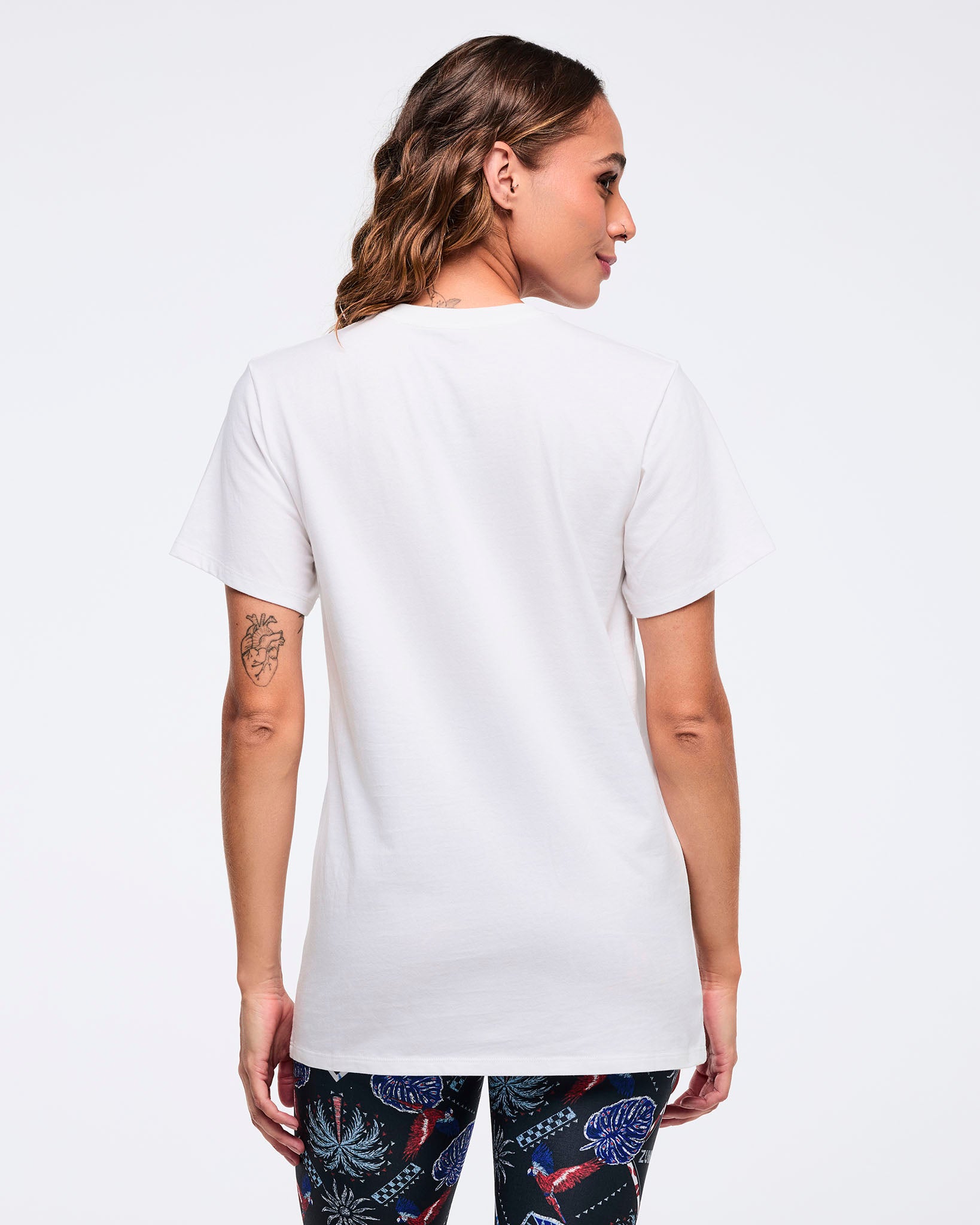 Back view of the white short-sleeve T-shirt on a female model, showing the plain back and straight hem, paired with dark tropical-print leggings.