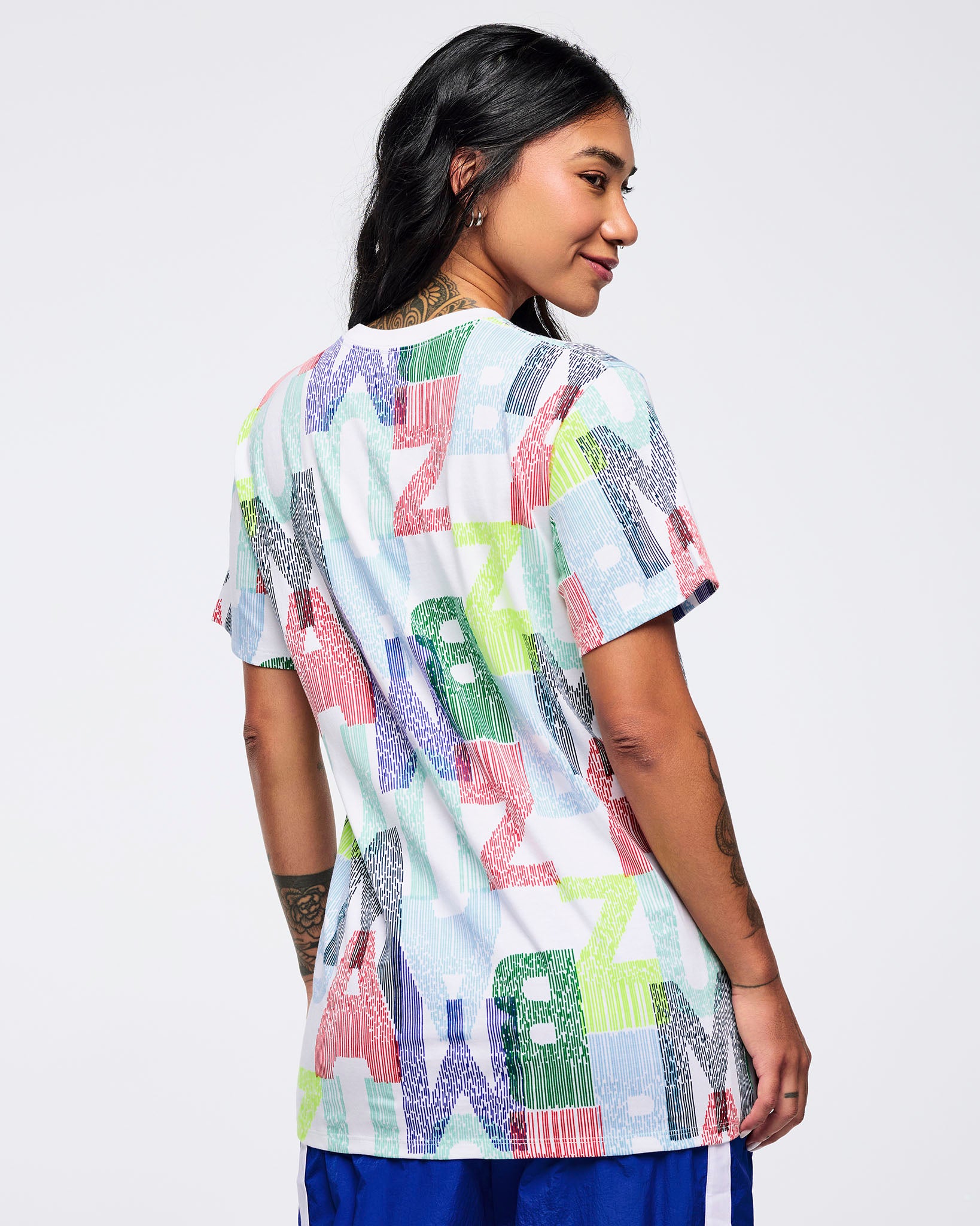 Back view of the all-over multicolor “ZUMBA” print T-shirt on a female model, showing the continuous graphic pattern and relaxed fit, paired with bright blue track pants with white side stripes.
