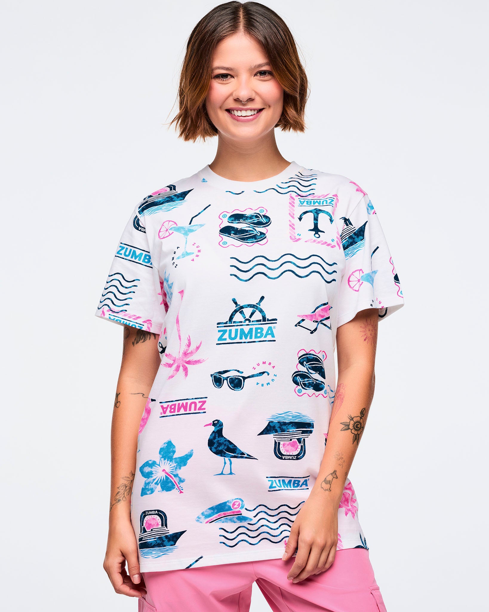 White short-sleeve crewneck Zumba graphic tee with an allover teal-and-pink beach print of waves, anchors, palm trees, sunglasses, boats, and logos; shown on a female model with pink pants.