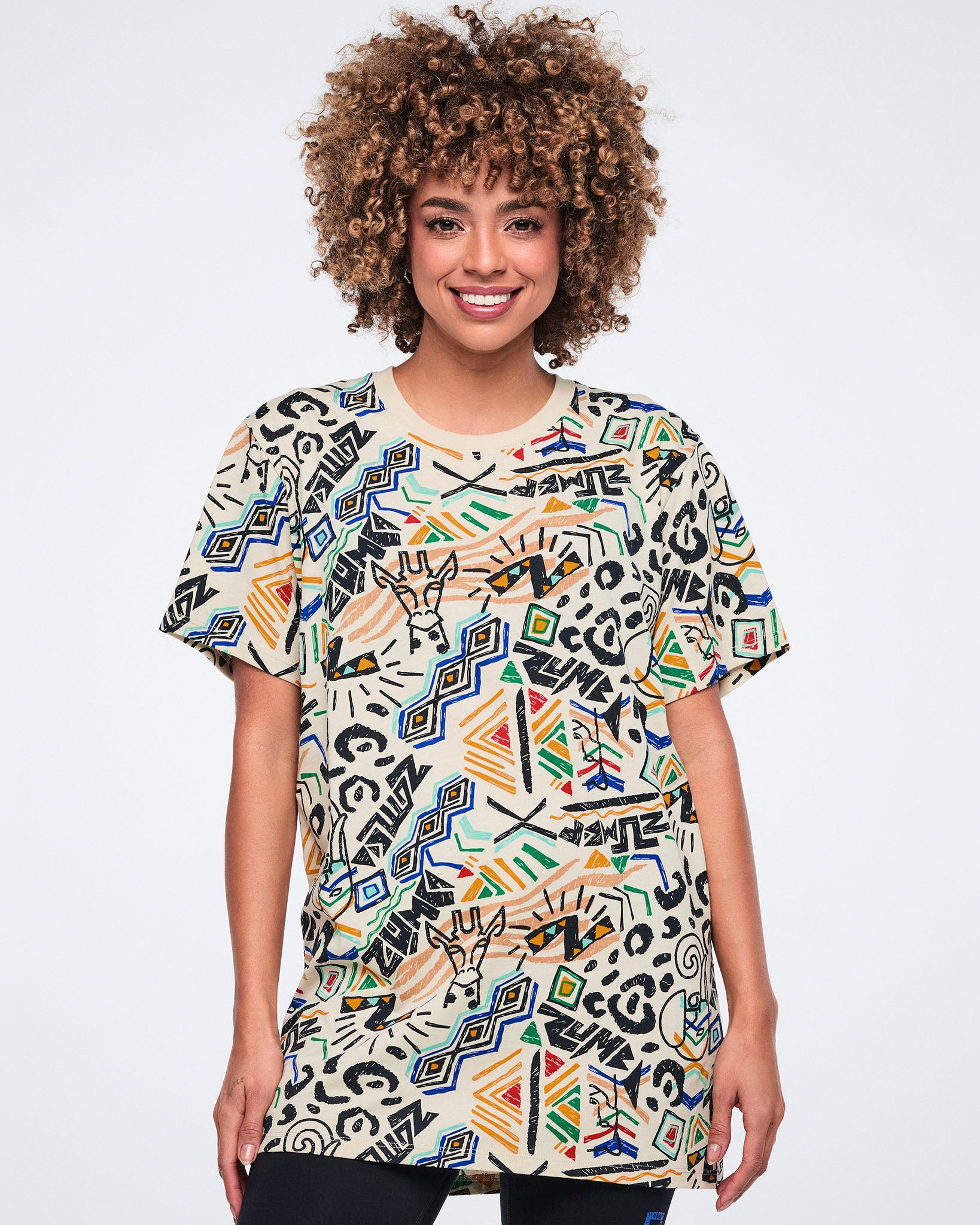 Cream short-sleeve graphic T-shirt with a crew neckline, oversized fit, and allover multicolor abstract print featuring geometric shapes, animal-inspired motifs, and Zumba logos. Female model wears the T-shirt over black leggings.