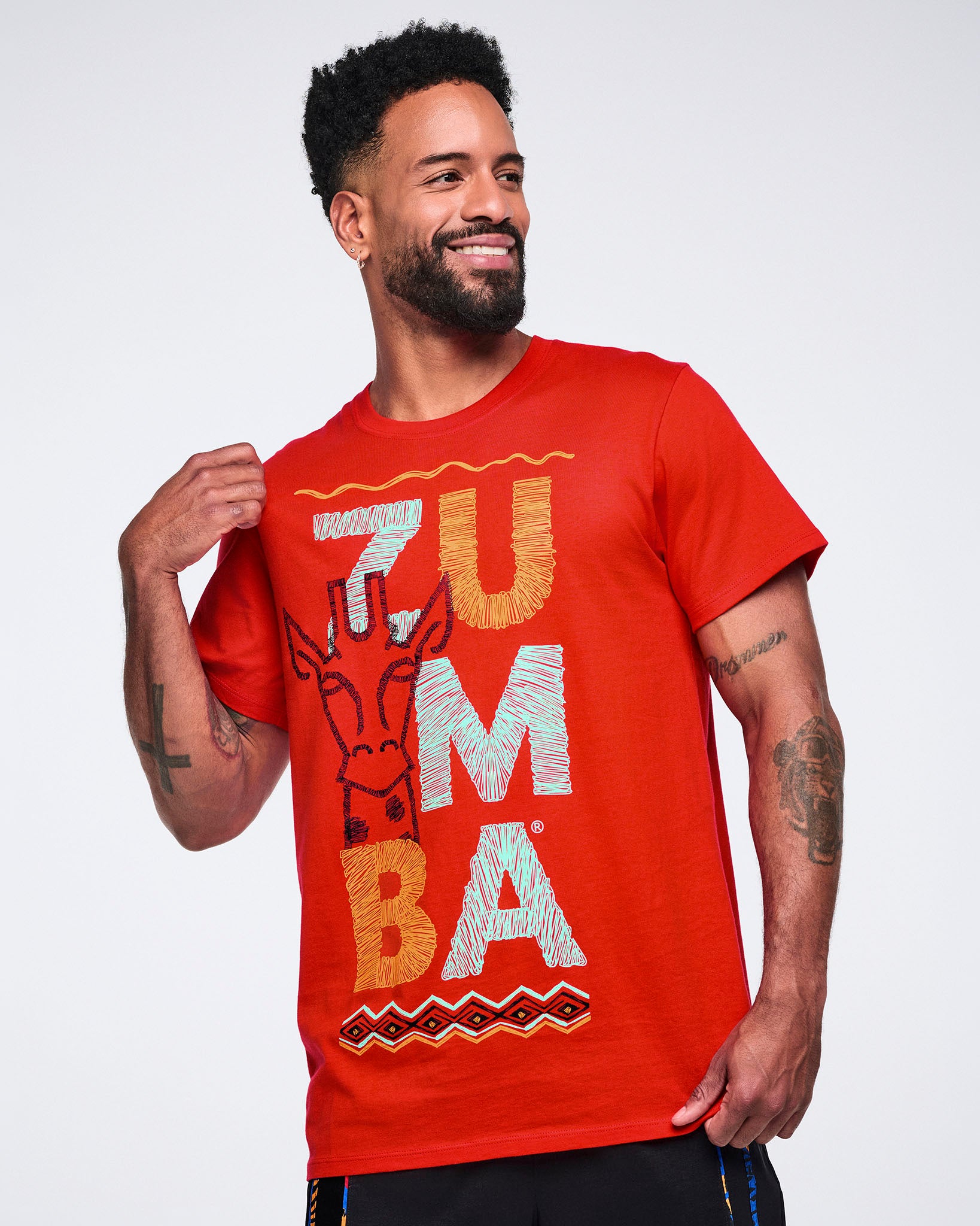 Male model wearing a red crewneck T-shirt with large multicolor Zumba lettering, a giraffe graphic, and a geometric border print on the front, paired with black athletic pants.