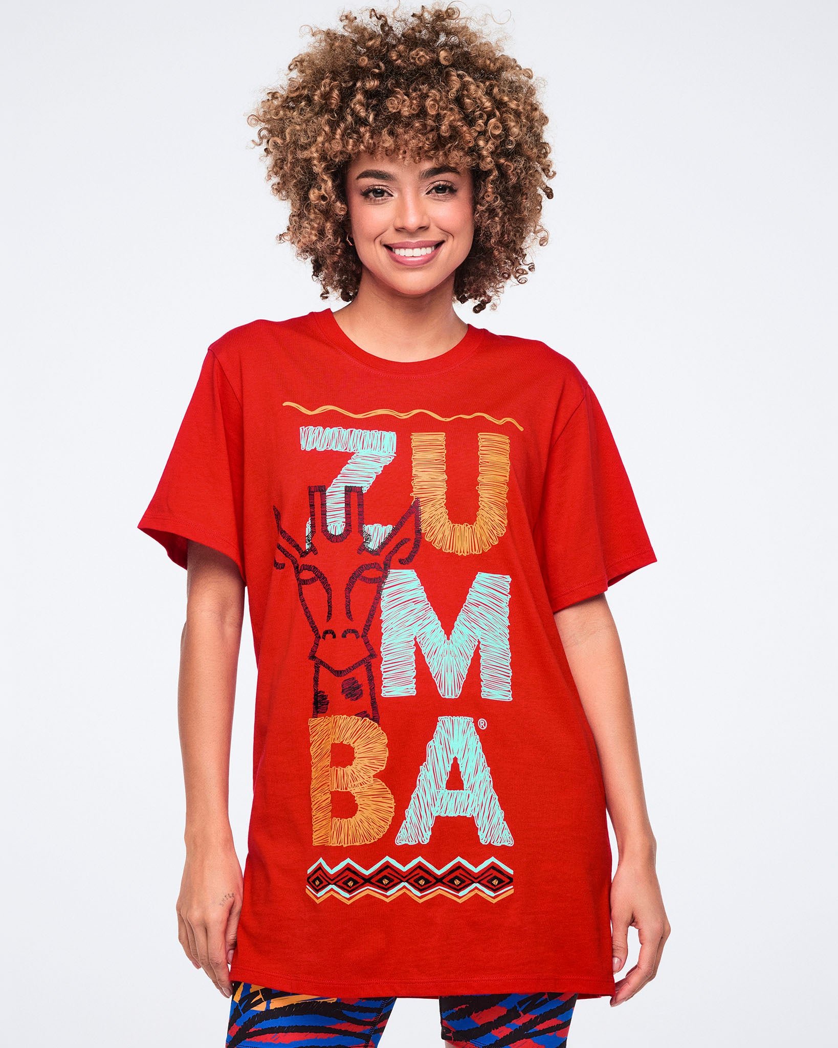 Female model wearing an oversized red crewneck T-shirt with large multicolor Zumba lettering, a giraffe graphic, and a geometric border print on the front, paired with multicolor zebra-print leggings.