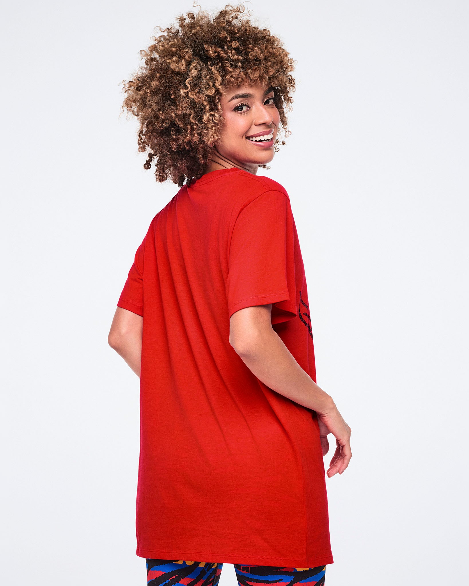 Female model wearing an oversized red crewneck T-shirt shown from the back with short sleeves and a relaxed fit, paired with multicolor zebra-print leggings.