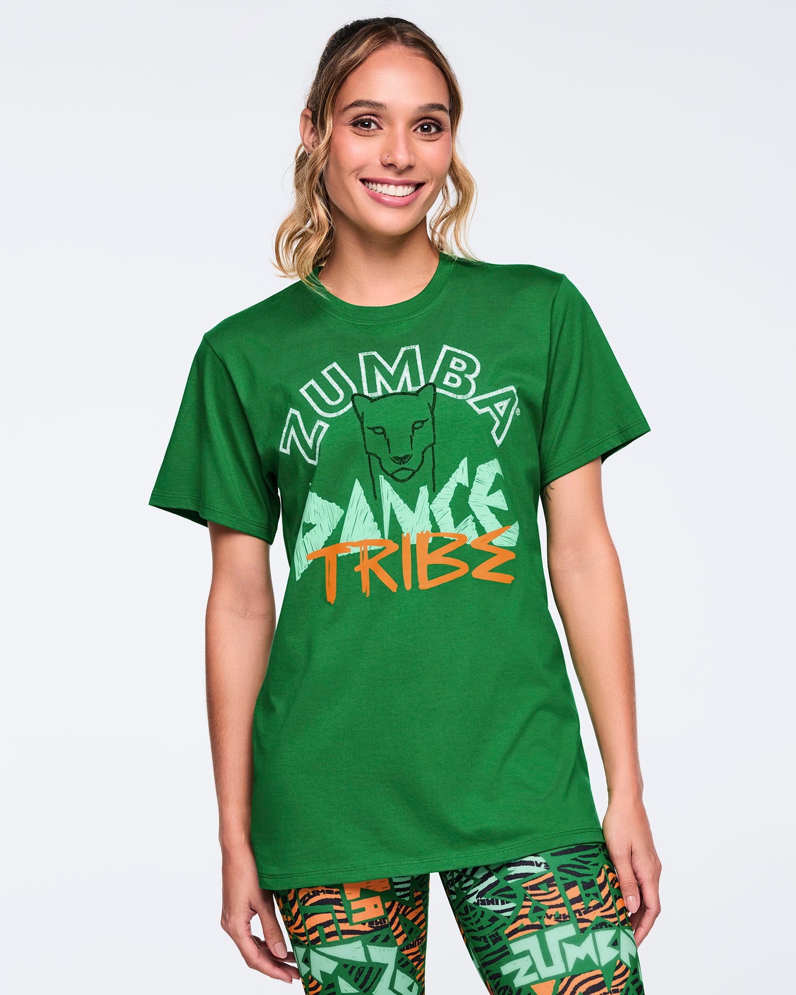 Green short-sleeve Zumba graphic T-shirt with a crew neckline and relaxed fit, featuring a front “Zumba Dance Tribe” design with a lion graphic in white, mint, and orange. Female model wears the T-shirt with green multicolor print leggings.