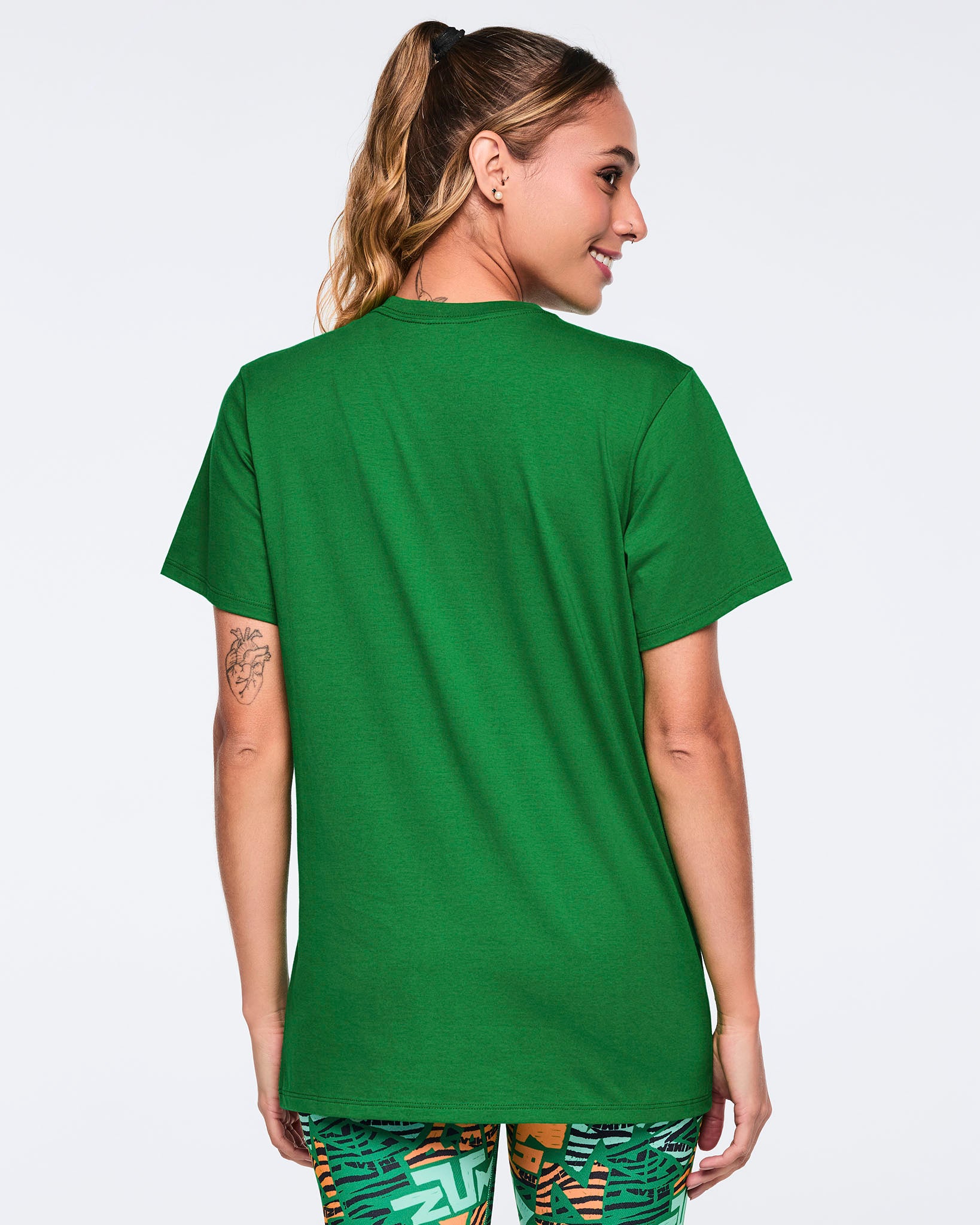 Back view of a green short-sleeve graphic T-shirt with a crew neckline, straight hem, and relaxed fit. Female model wears the T-shirt with green multicolor print leggings.