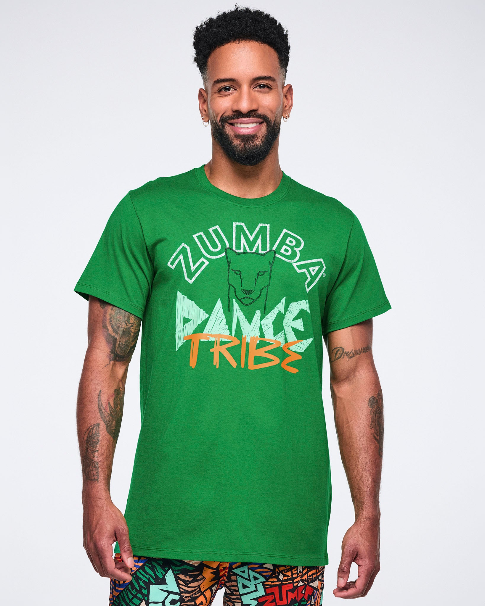 Green short-sleeve Zumba graphic T-shirt with a crew neckline and relaxed fit, featuring a front “Zumba Dance Tribe” design with a lion graphic in white, mint, and orange. Male model wears the T-shirt with multicolor print pants.