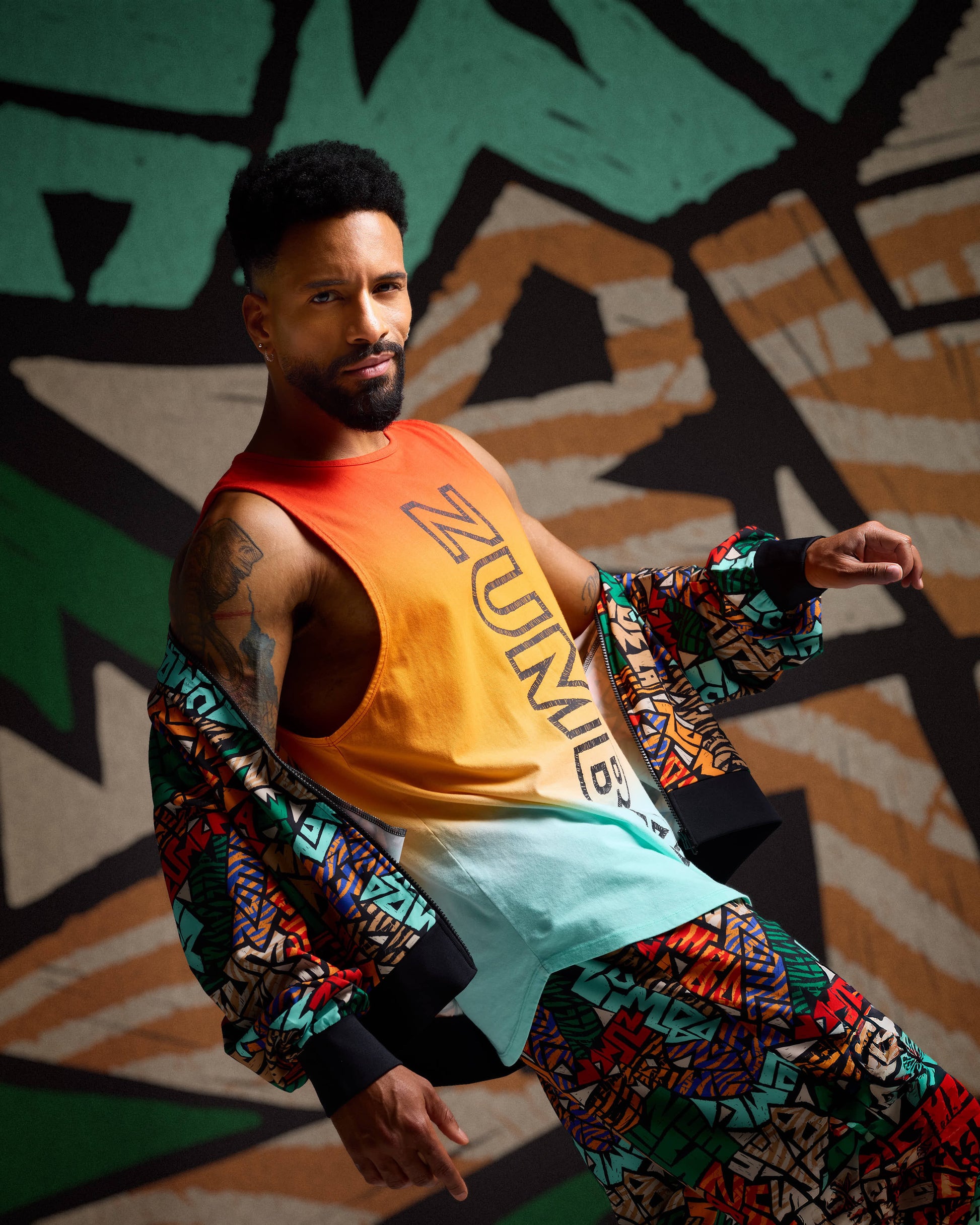 Male model wearing a sleeveless longline tank in a red-to-orange-to-mint gradient with large vertical black Zumba lettering, layered under a multicolor abstract-print jacket and matching pants against a colorful background.