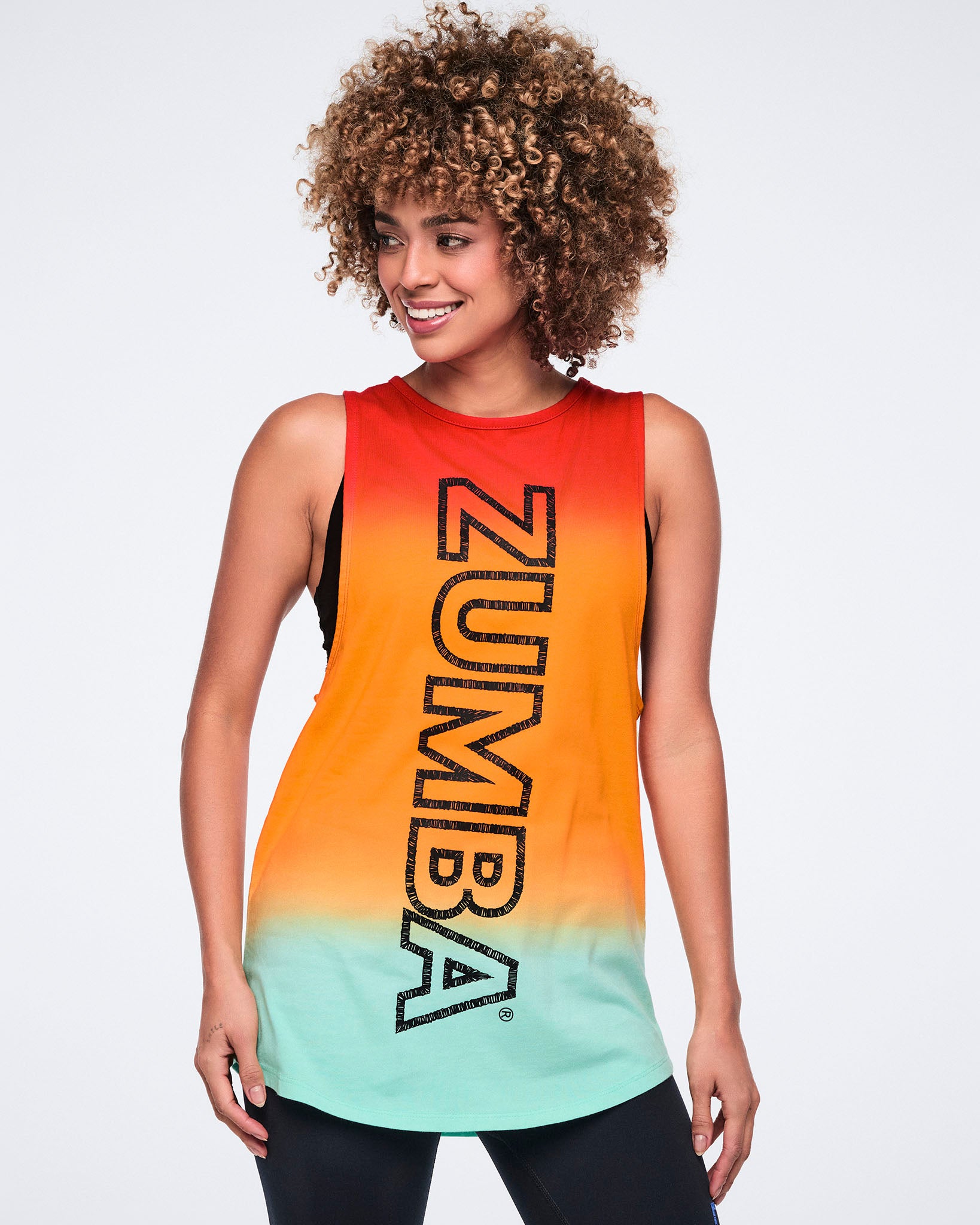 Female model wearing a sleeveless longline tank in a red-to-orange-to-mint gradient with large vertical black Zumba lettering, paired with black leggings.