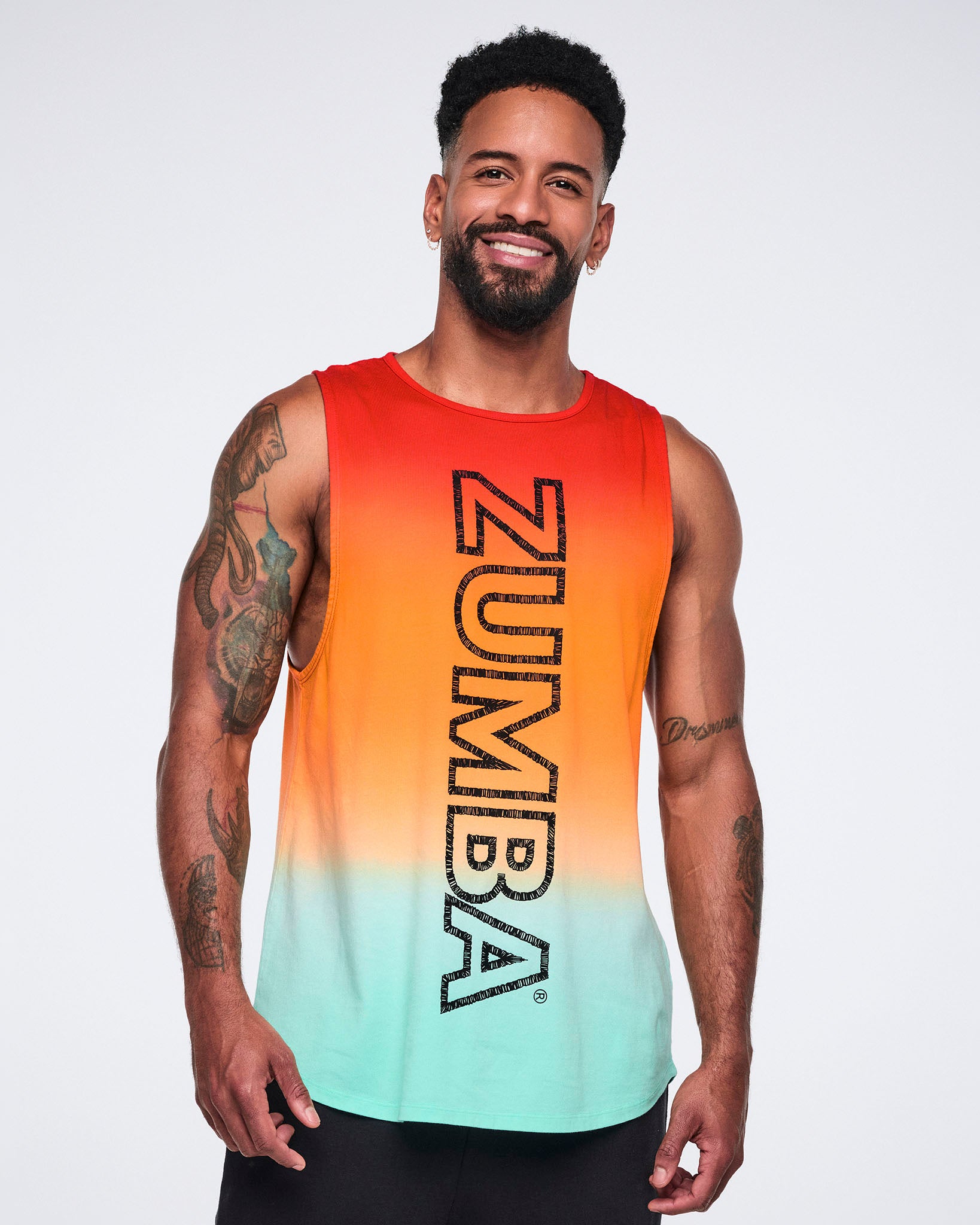 Male model wearing a sleeveless longline tank in a red-to-orange-to-mint gradient with large vertical black Zumba lettering, paired with black pants.
