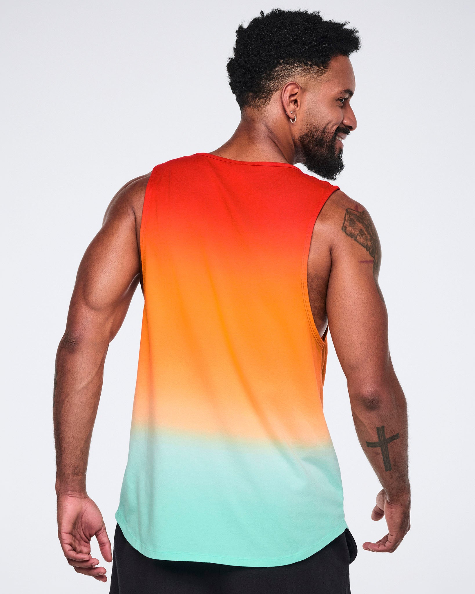 Male model wearing a sleeveless longline tank shown from the back in a red-to-orange-to-mint gradient with a relaxed fit and curved hem, paired with black pants.