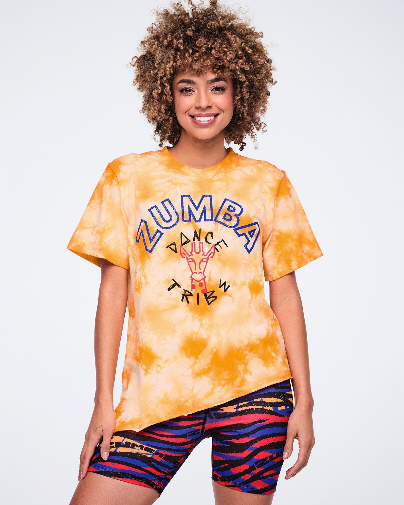 Female model wearing an oversized orange and white tie-dye T-shirt with blue Zumba text and a “Dance Tribe” giraffe graphic, paired with multicolor zebra-print biker shorts.