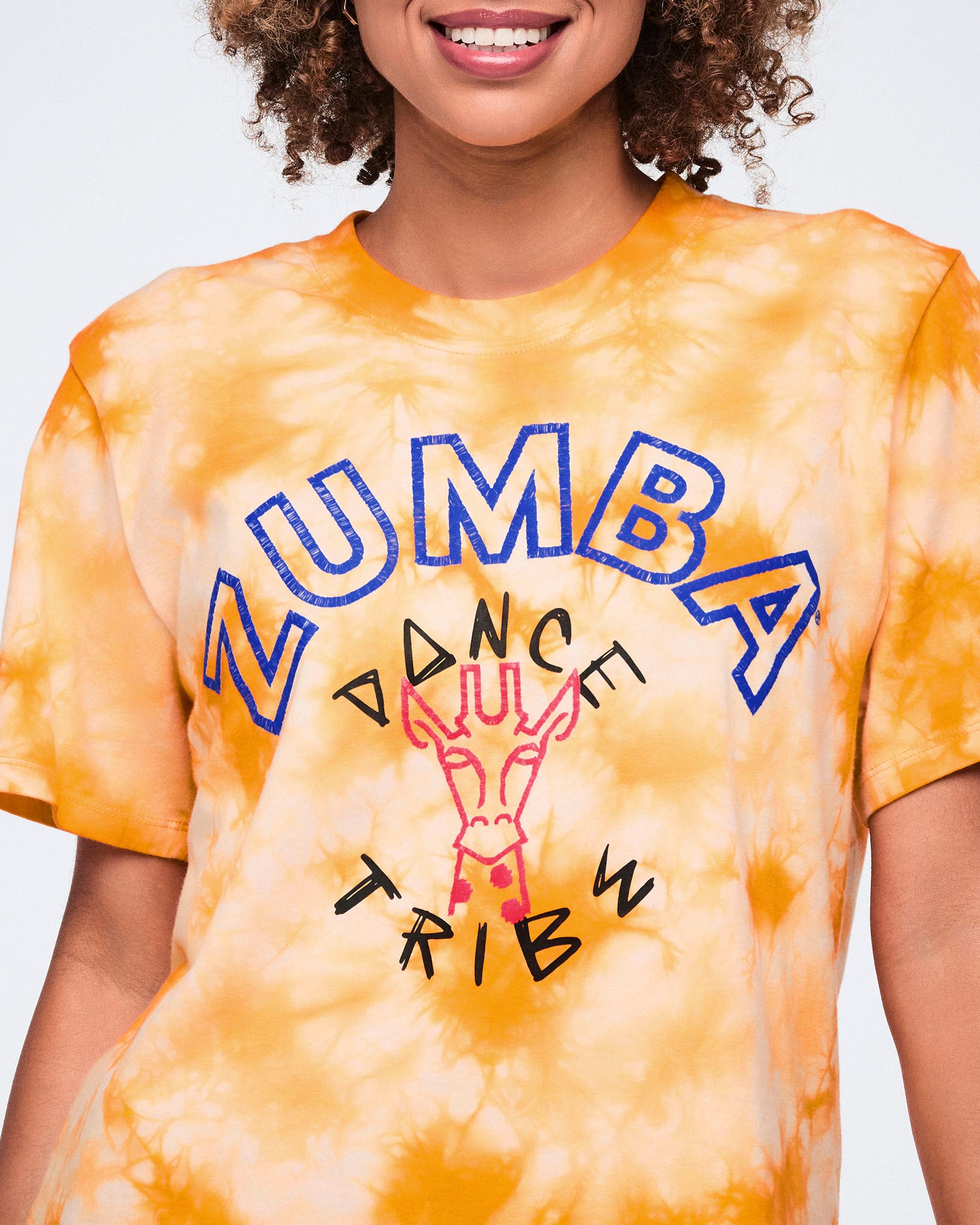 Close-up of an oversized orange and white tie-dye T-shirt with blue Zumba text, black “Dance Tribe” lettering, and a pink giraffe graphic on the front.
