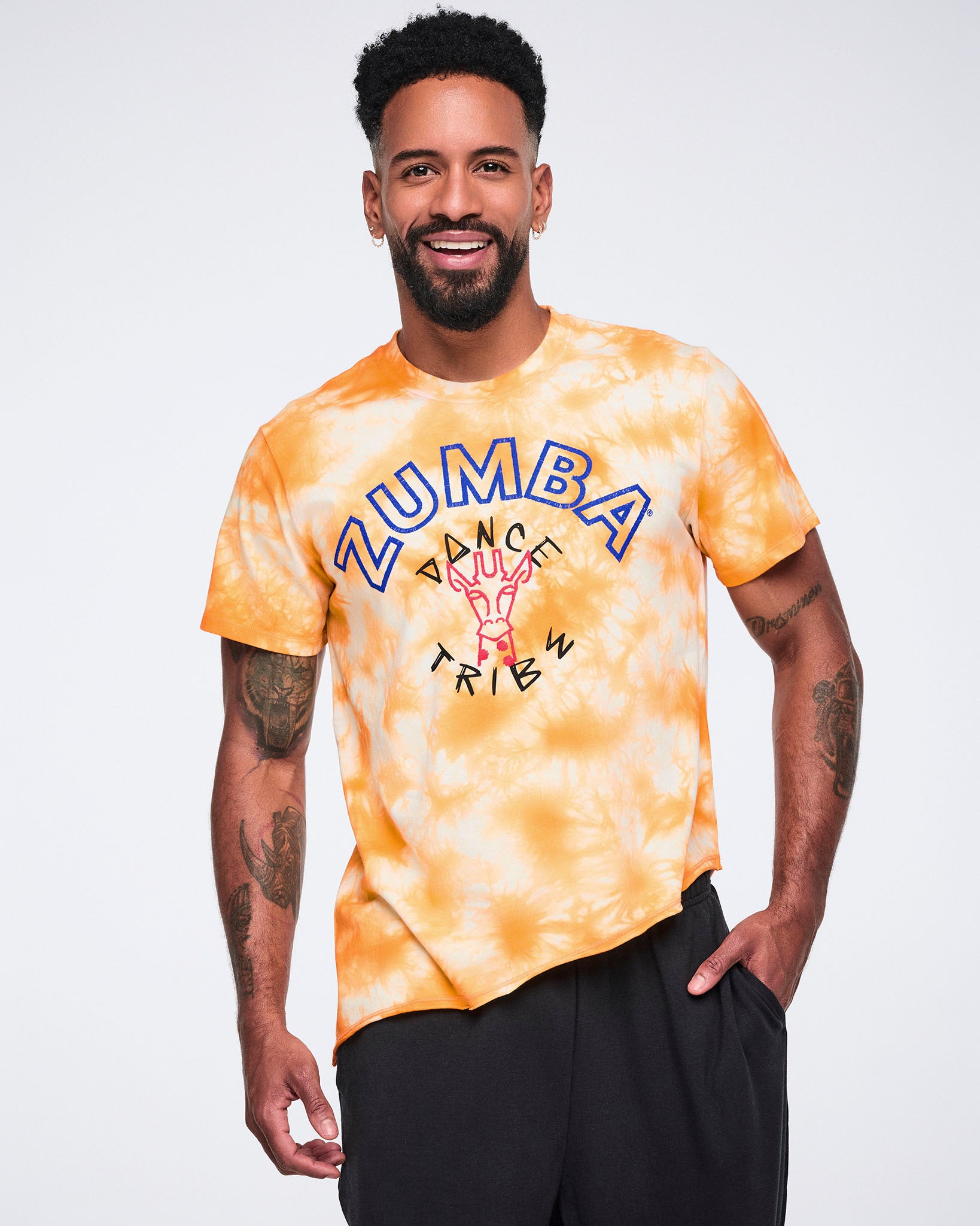 Male model wearing an oversized orange and white tie-dye T-shirt with blue Zumba text and a “Dance Tribe” giraffe graphic, paired with black wide-leg athletic pants.