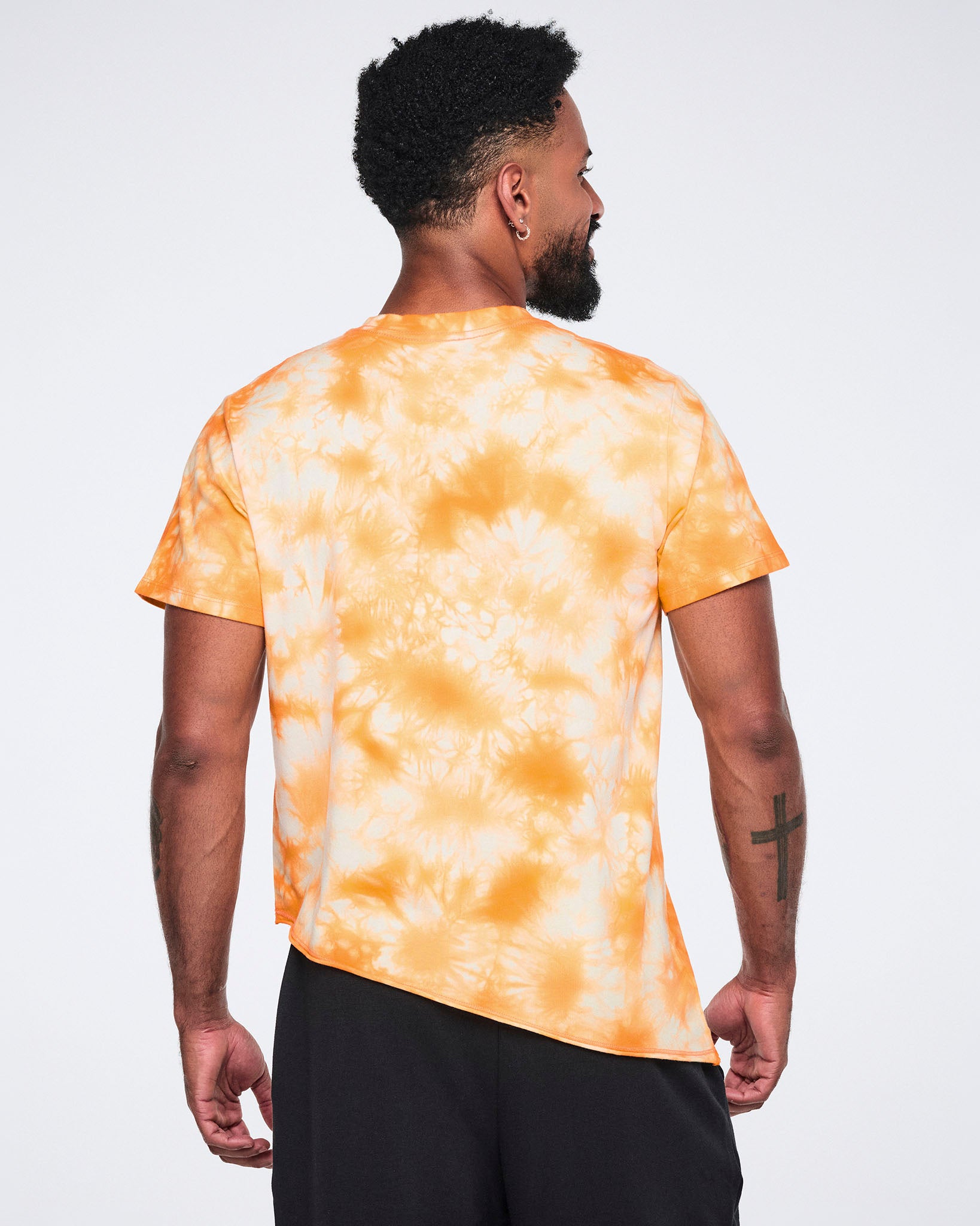 Male model wearing an oversized orange and white tie-dye T-shirt shown from the back with short sleeves, an asymmetric hem, and black wide-leg athletic pants.