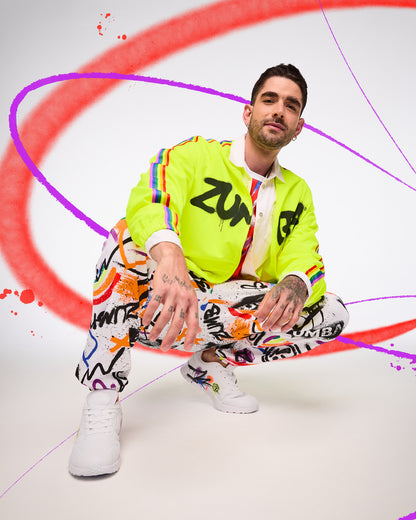Male model wearing a bright yellow jacket with 'Zumba' black text, colorful pants, and white sneakers on a white background with red and purple abstract designs.