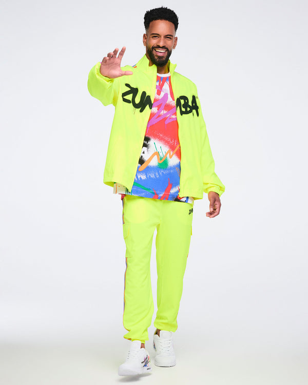 Male model wearing a bright yellow jacket over an abstract graphic T‑shirt, paired with bright yellow pants and white Zumba sneakers on a white background.