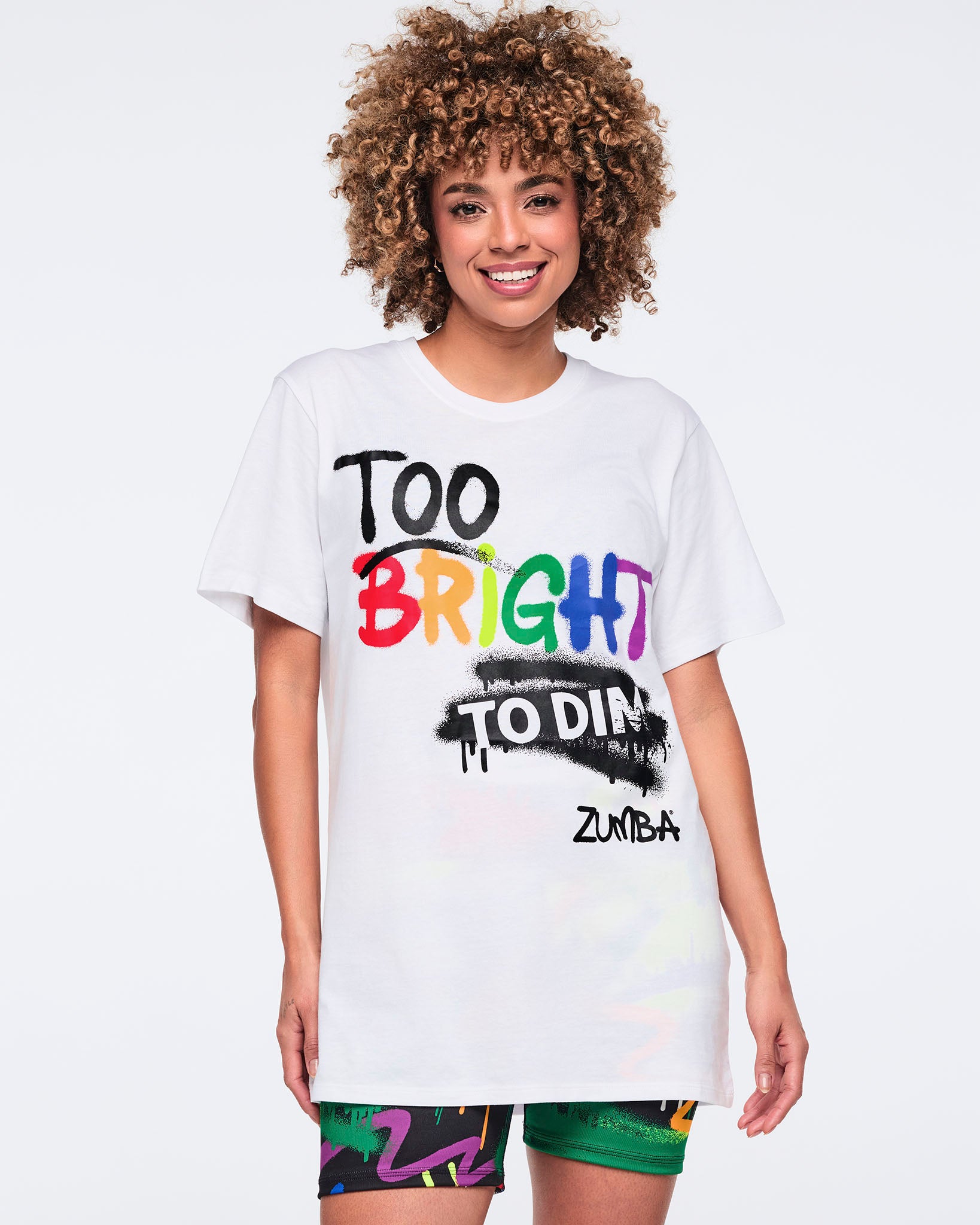 Female model wearing a white T‑shirt that reads “Too bright to dim” in mostly black text with “bright” in rainbow colors, paired with colorful biker shorts on a white background.