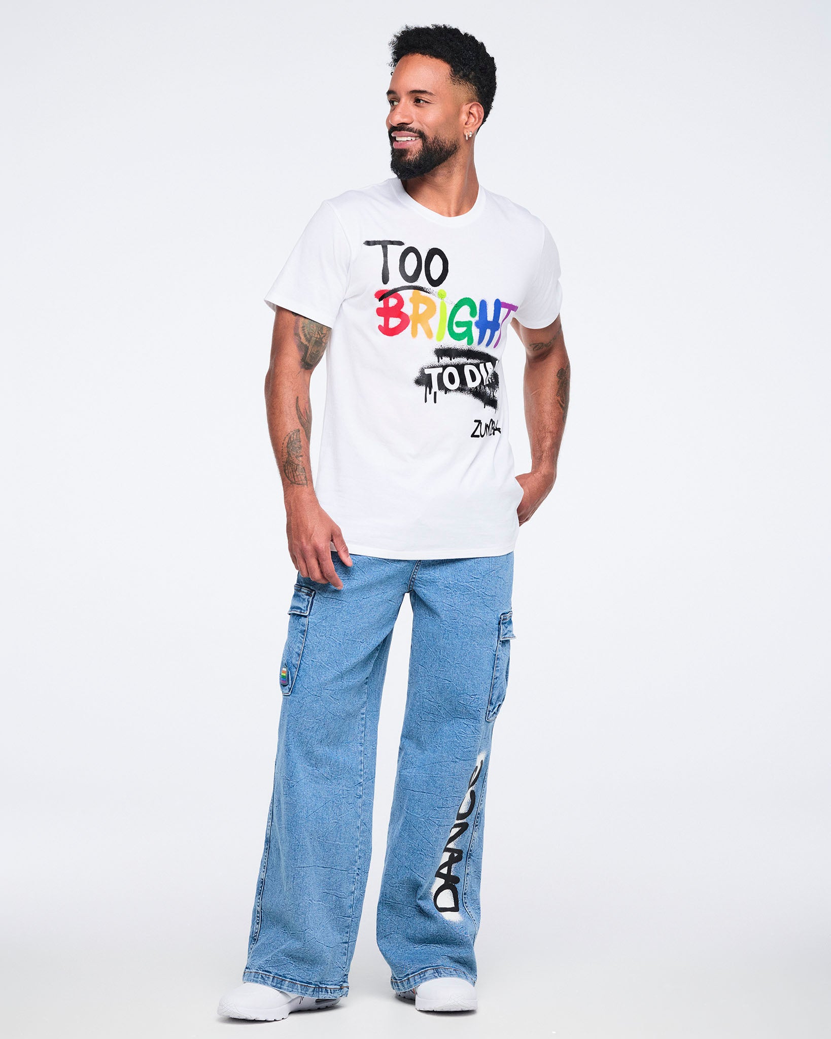 Male model wearing a white T‑shirt with “Too bright to dim Zumba” mostly in black text except “bright” in rainbow colors, paired with denim cargo pants on a white background.