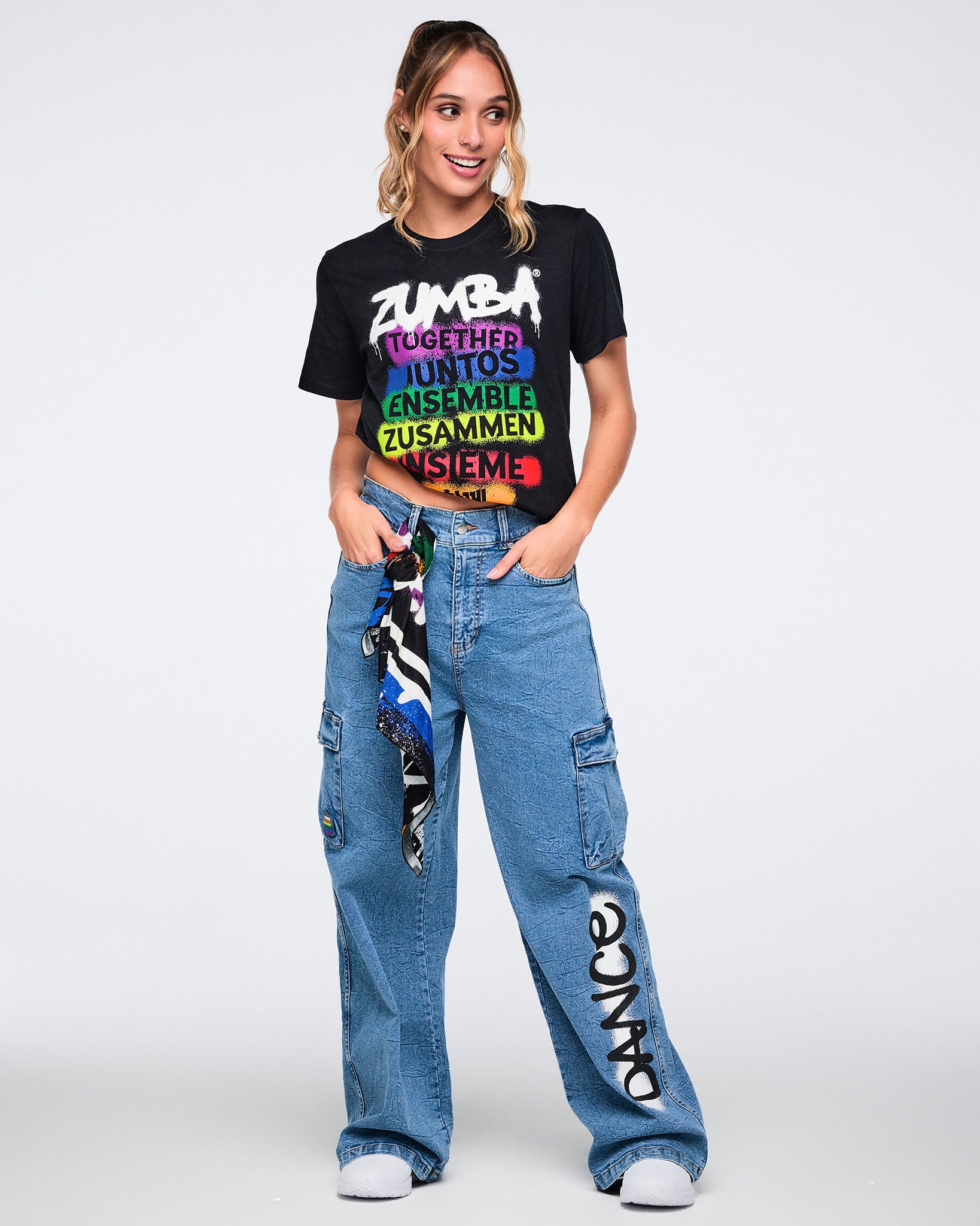 Female model wearing a black Zumba t-shirt with colorful text and denim cargo pants on a white background.