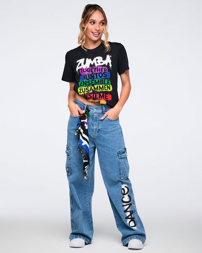 Female model wearing a black Zumba t-shirt with colorful text and denim cargo pants on a white background.