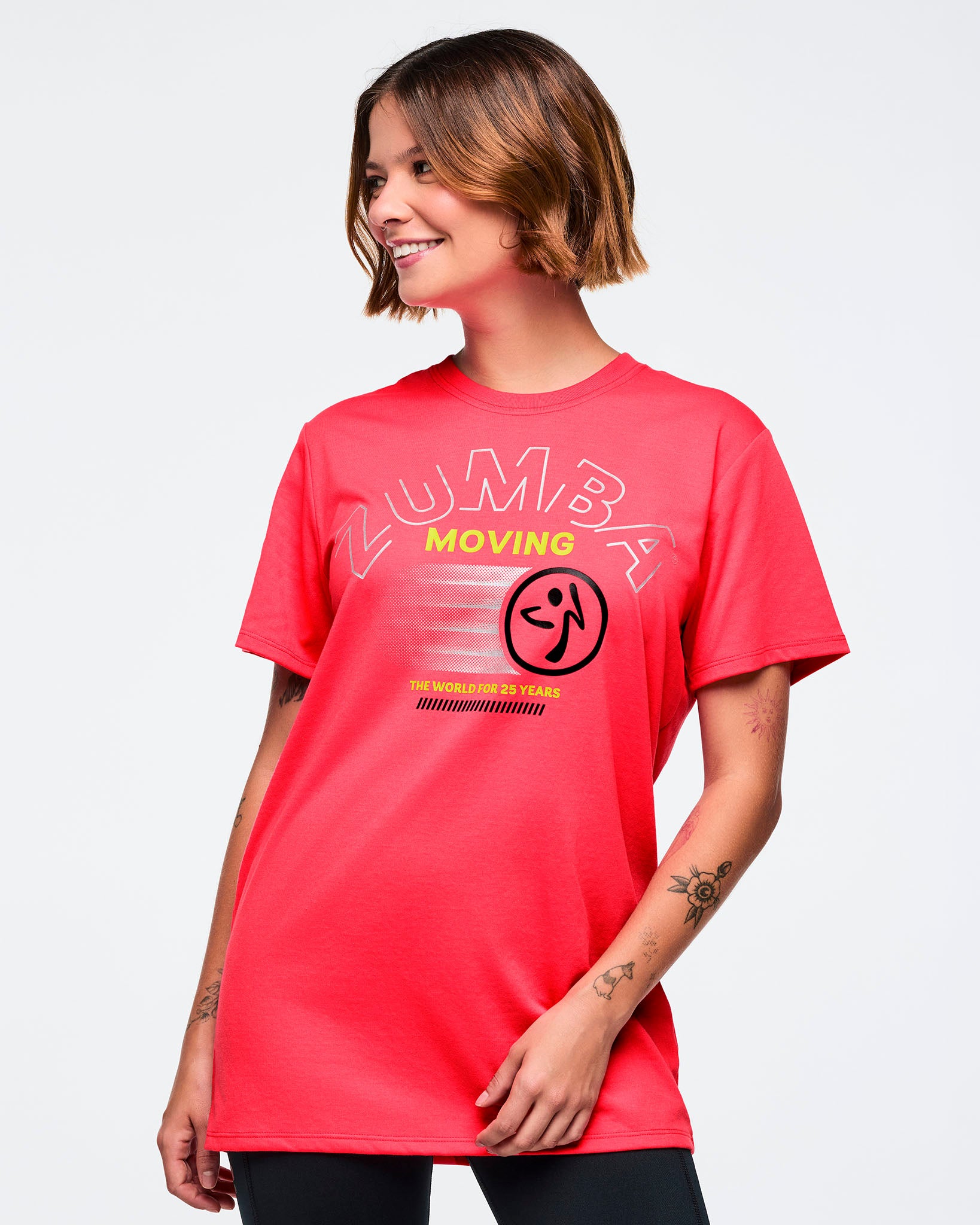 Bright coral pink Zumba T-shirt with a large tonal ZUMBA graphic, yellow “MOVING” text, and black Zumba logo on the front. Female model is smiling and wearing the short-sleeve crewneck tee with black leggings.