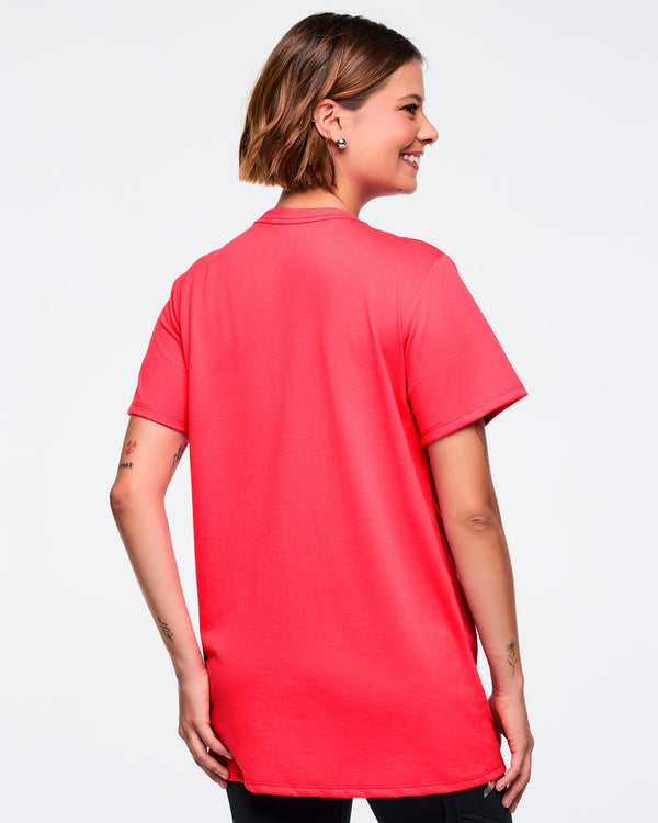 Bright coral pink Zumba T-shirt shown from the back with a clean, plain design. Female model is turned sideways and wearing the short-sleeve crewneck tee with black leggings.