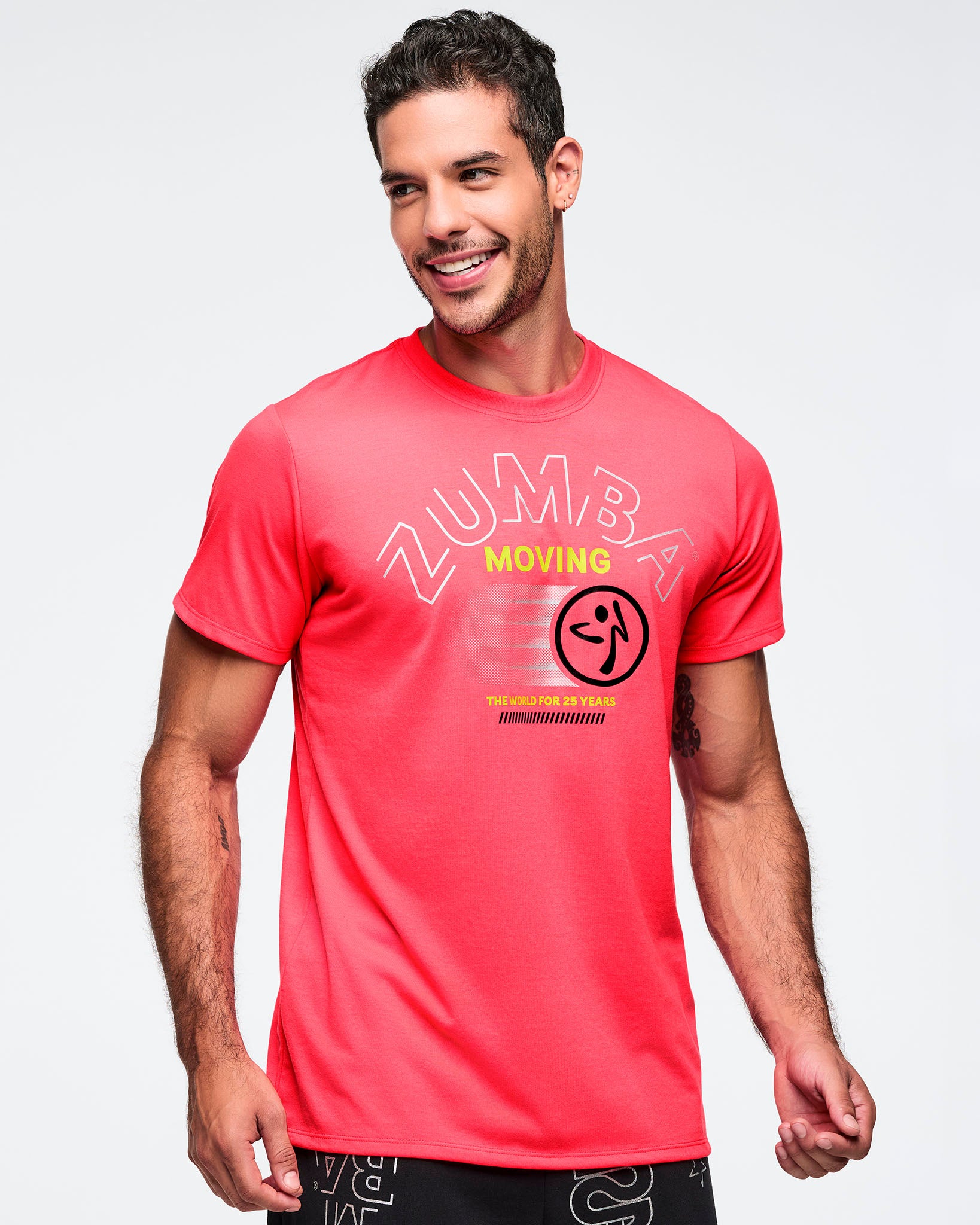 Bright coral pink Zumba T-shirt with a large tonal ZUMBA graphic, yellow “MOVING” text, and black Zumba logo on the front. Male model is smiling and wearing the fitted short-sleeve crewneck tee with black graphic pants.