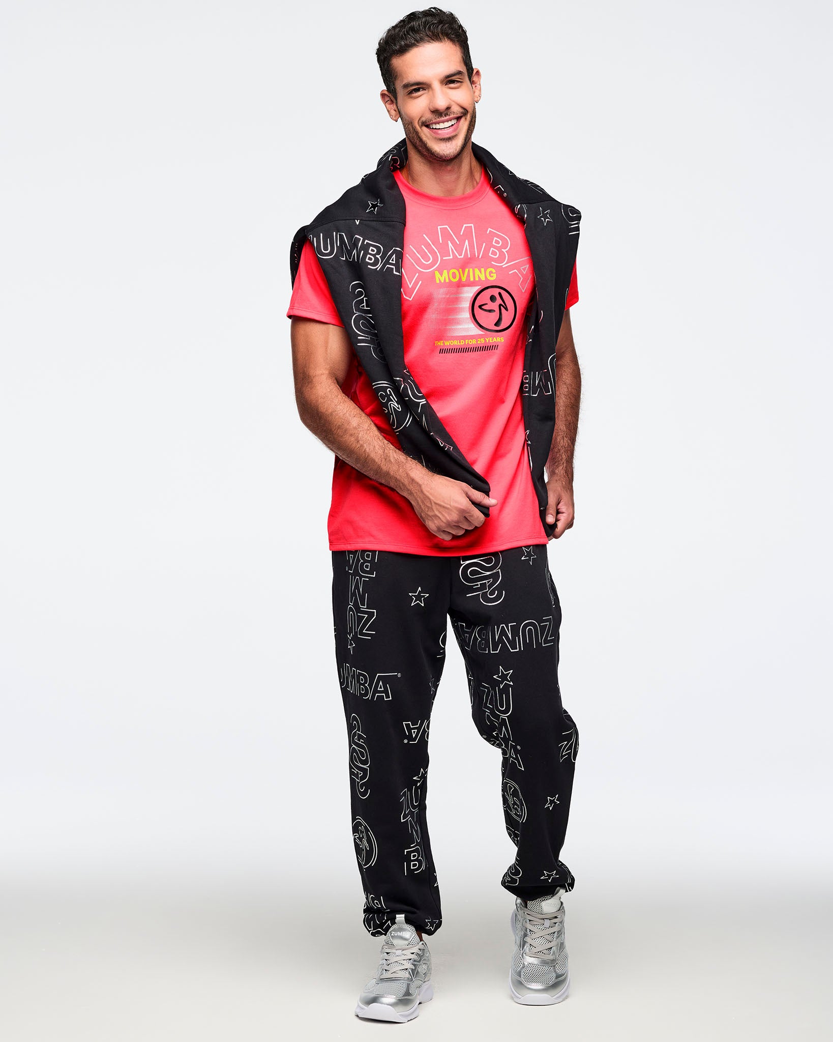 Bright coral pink Zumba T-shirt with a large tonal ZUMBA graphic, yellow “MOVING” text, and black Zumba logo on the front. Male model is wearing the short-sleeve crewneck tee with black printed joggers, silver sneakers, and a black printed sweatshirt draped over his shoulders.