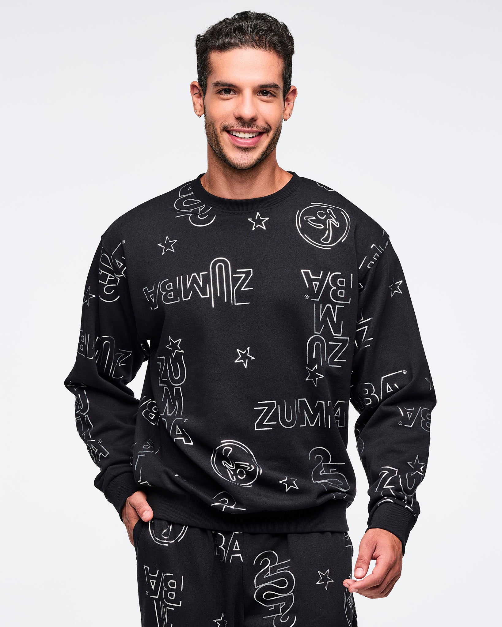 Black Zumba sweatshirt with an allover silver logo, star, and icon print. Male model is smiling and wearing the long-sleeve crewneck sweatshirt with matching black printed joggers.