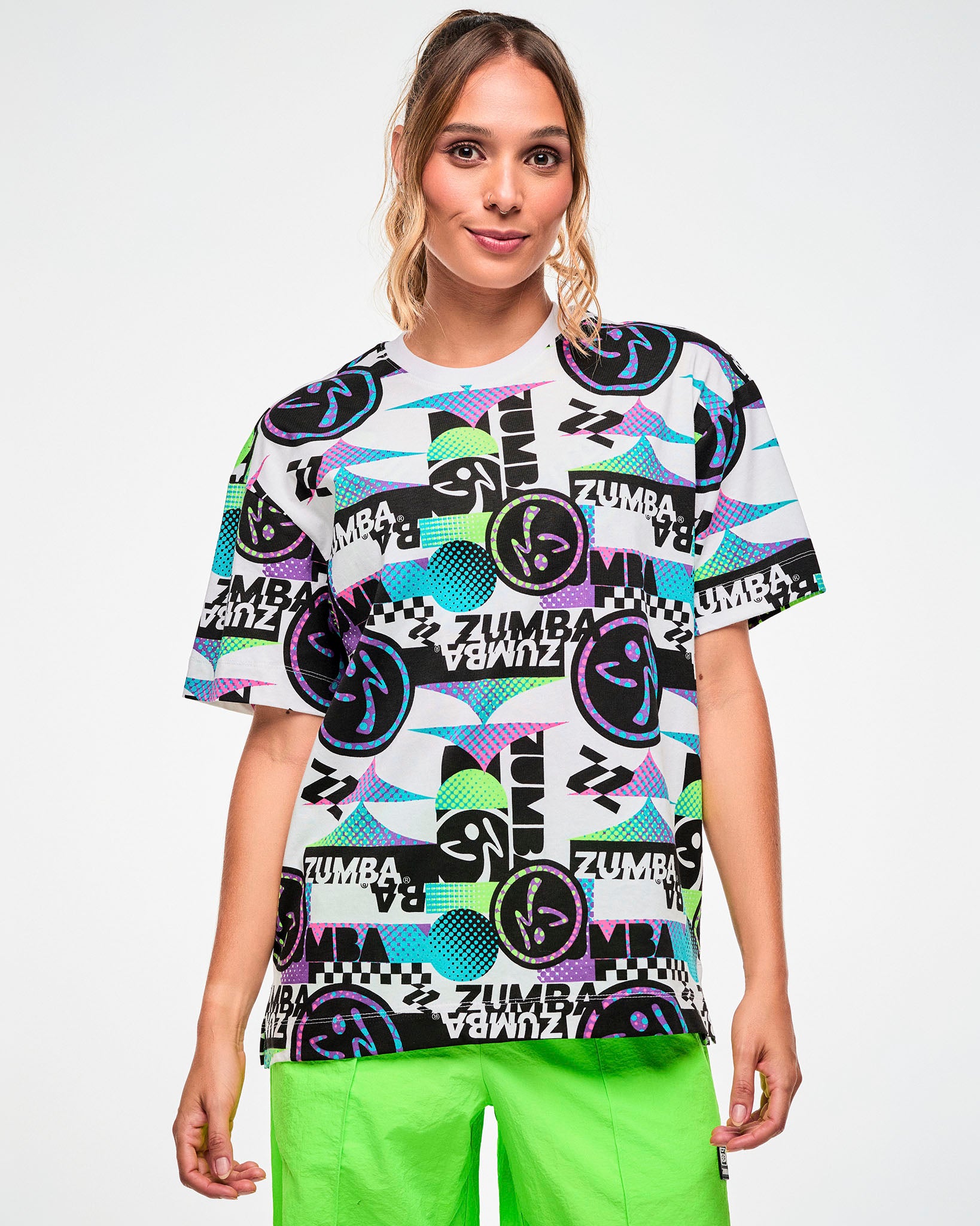 Female model wearing a white tee with a busy graphic pattern featuring "ZUMBA" text, Zumbito logos, and other patterns, paired with green pants.