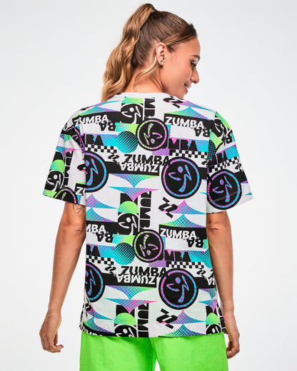 Back view of a female model wearing a white tee with a busy graphic pattern of repeated "ZUMBA" text and Zumbito logos.
