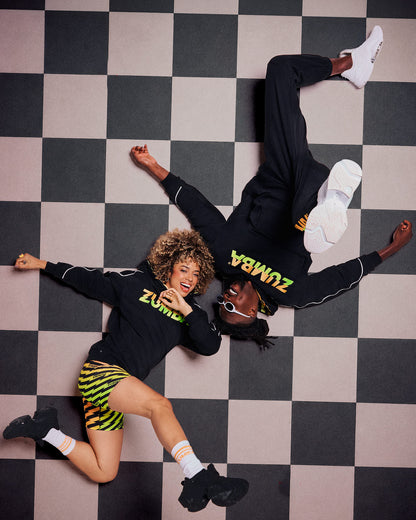 Male and female models wearing black hoodies with a gradient "ZUMBA" logo and sneakers, one paired with lightning bolt-print biker shorts, lying down on a black and white checkered floor posing.