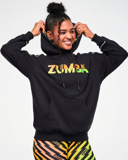 Female model wearing a black ZUMBA hoodie with an orange, yellow, and green ombre lightning bolt-print, paired with matching biker shorts.