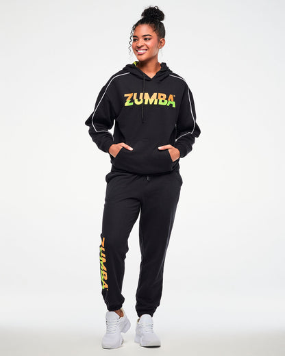 Female model wearing a black hoodie and matching joggers with a gradient "ZUMBA" logo, paired with white sneakers.