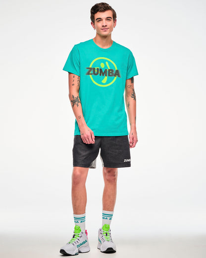 Male model wearing an aqua tee with a yellow Zumbito logo and halftone black dot "ZUMBA" text, paired with black shorts and white sneakers.