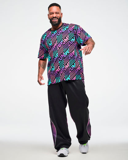 Male model wearing a black tee with a pink, aqua, and purple diagonal striped "ZUMBA" broken patterned print, paired with black joggers featuring purple side panels.