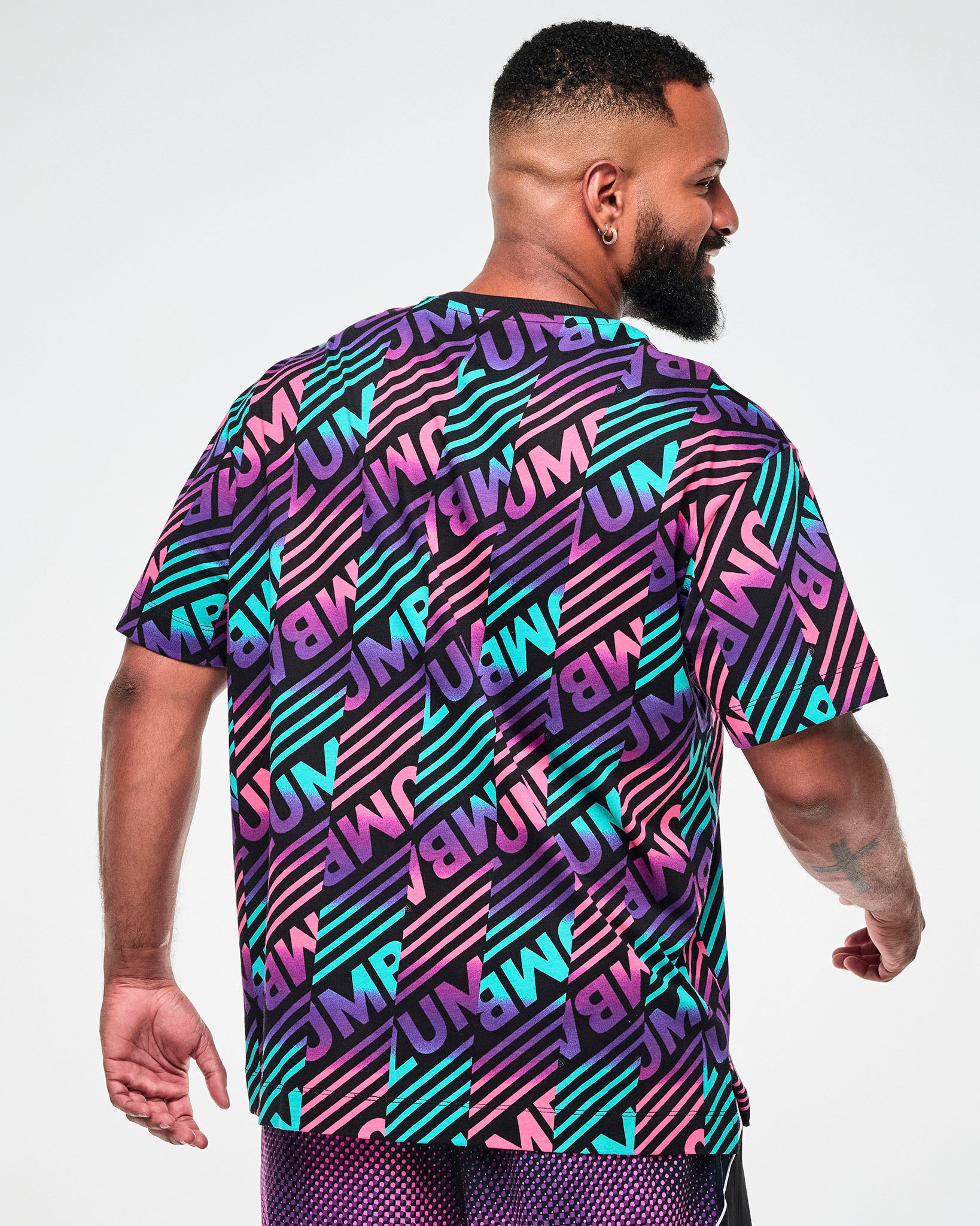 Shown from the back, male model wearing a black tee with a colorful pink, aqua, and  purple diagonal striped "ZUMBA" broken patterned print.