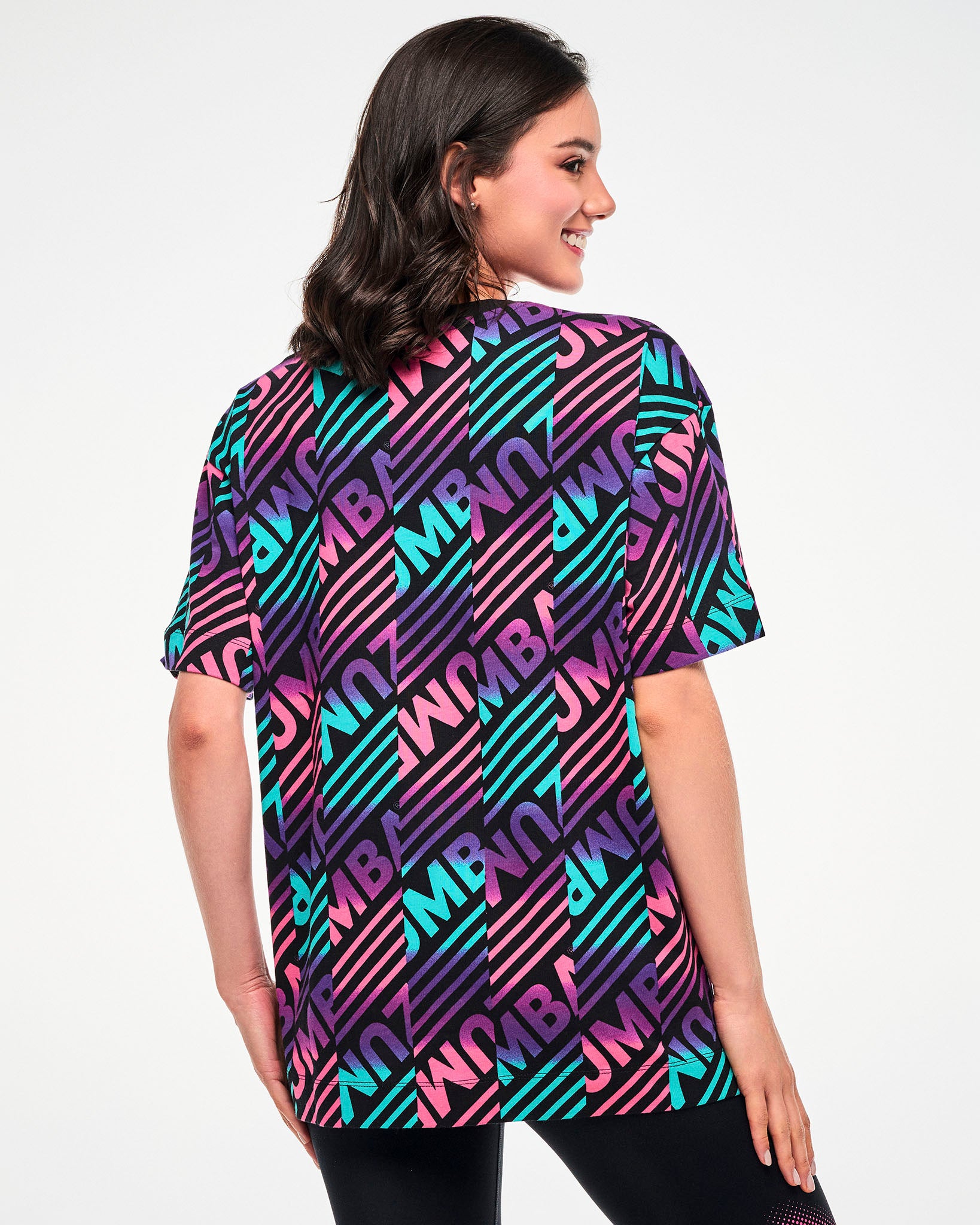 Shown from the back, female model wearing a black tee with a colorful pink, aqua, and purple diagonal striped "ZUMBA" broken patterned print.