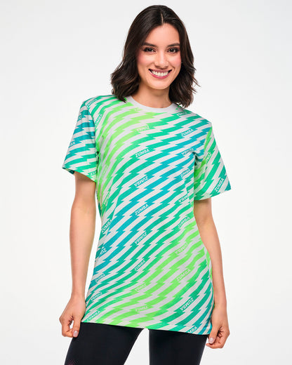 Female model wearing a green and aqua lightning bolt-striped tee with repeated "ZUMBA" text and paired with black leggings.